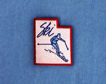 May include: A red and white embroidered patch with a skier in blue and white clothing. The patch says "Ski" in blue cursive lettering.