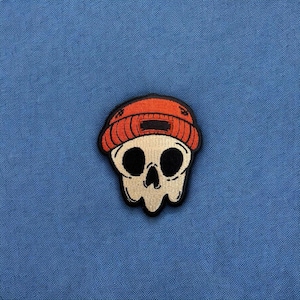 May include: Embroidered patch featuring a stylized skull wearing an orange beanie. The skull has large, black eye sockets and a small nose cavity. The patch is outlined in black and set against a blue background.