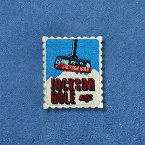 May include: Embroidered patch featuring a ski lift and the words "Jackson Hole" in red and white. The patch is in a postage stamp shape with a blue background and white border. The patch is on a blue fabric background.
