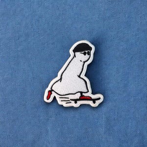 May include: White embroidered patch of a cartoon character wearing a black hat and sunglasses, riding a skateboard with red wheels.