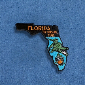May include: An embroidered patch shaped like the state of Florida. The top section is black with orange text reading FLORIDA THE SUNSHINE STATE. The bottom section is light blue featuring a green alligator and an orange fruit with white blossoms.