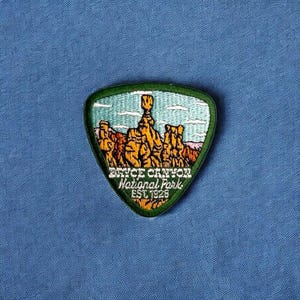 May include: Green, triangular patch with an embroidered image of Bryce Canyon National Park. The image shows a group of hoodoos, which are tall, thin rock formations. The text "Bryce Canyon National Park Est. 1928" is embroidered on the patch.