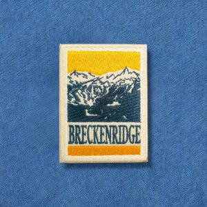May include: Embroidered patch featuring a mountain range scene with the word "BRECKENRIDGE" in white lettering. The patch has a yellow sky, a blue mountain range, and a cream-colored border, set against a blue fabric background.