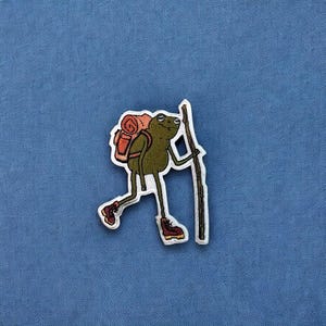 May include: A green frog with a backpack and a stick in its hand. The frog is wearing brown boots and has a red and brown backpack.