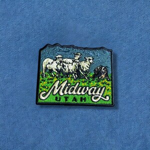 May include: Embroidered patch featuring a scenic view of Midway, Utah. The patch depicts sheep grazing in a field with the town's name in a green, cursive font. The background is blue.