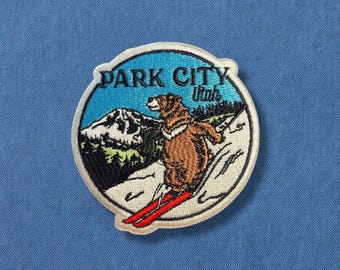 May include: Embroidered patch with a blue background and white border. The patch features a brown bear skiing down a snowy mountain with the text "PARK CITY Utah" in white.