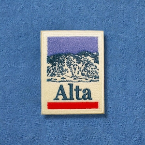 May include: Embroidered patch featuring a mountain landscape with the word "Alta" in navy blue. The patch has a red horizontal stripe at the bottom and a purple sky. The patch is rectangular with a white border, set against a blue fabric background.