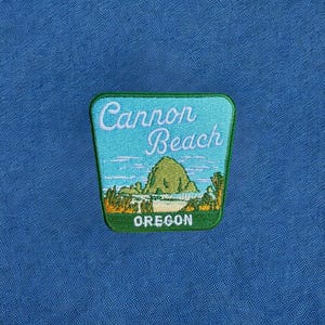 May include: A blue fabric background featuring a rectangular embroidered patch with rounded corners. The patch depicts a coastal scene with a large green rock formation, white text reading Cannon Beach, and the word OREGON in white at the bottom.
