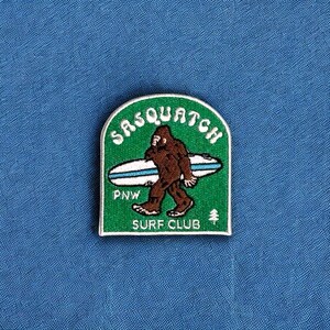 May include: Green embroidered patch with a white outline. The patch features a cartoon Sasquatch holding a surfboard with the words "Sasquatch Surf Club" and "PNW" on it.