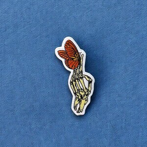 May include: A skeleton hand holding a butterfly with orange and yellow wings. The butterfly is in the hand's palm and the hand is reaching out.
