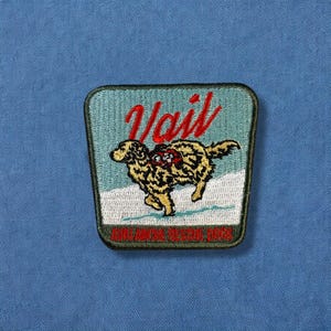 May include: Embroidered patch featuring a running dog in yellow and black, set against a snowy landscape and a teal background. The word "Vail" is written in red script above the dog. The bottom text reads "AVALANCHE RESCUE DOGS".