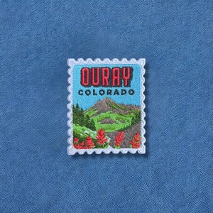 May include: Embroidered patch featuring a scenic view of a mountain landscape with the text "OURAY COLORADO" in red. The patch has a light blue background and a white scalloped border, resembling a postage stamp.
