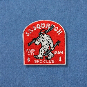 May include: Red embroidered patch with a white sasquatch holding skis, the text "Sasquatch" above and "Park City Ski Club" below, and the year "1869" to the right.