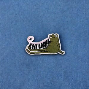 May include: Green frog with a pink tongue sticking out, lying on its back with its legs in the air. The frog is wearing a white patch with the text "EAT LOCAL" in black letters.
