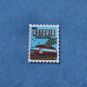 May include: Embroidered patch in the shape of a postage stamp. The patch features a red and white trolley car, a green bridge, and the words "Visit Astoria Oregon" in white and black text. The patch has a white border and is set against a blue background.