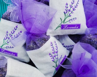 May include: Purple lavender sachets with a white fabric and lavender embroidery. The sachets are tied with a purple ribbon and have the word "Lavender" printed on them.