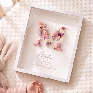 May include: A white shadow box frame containing a letter M formed from dried flowers in shades of pink, purple, and cream. Below the letter, the text reads Mila, 28 October 2025, and Long waited for and much loved, printed in a dark grey font.