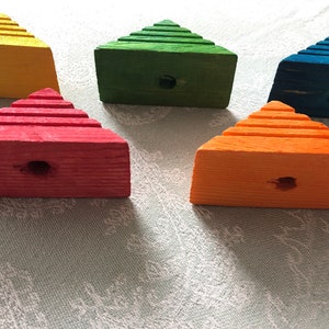 Puede incluir: Un conjunto de cinco bloques de madera coloridos en forma de pirámide escalonada. Cada bloque está pintado de un color diferente: amarillo, verde, rojo, naranja y azul. Cada bloque tiene un agujero perforado en la parte delantera. Dispuestos sobre una superficie verde claro.