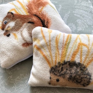 Puede incluir: Dos almohadas de felpa blanca con imágenes bordadas de una ardilla y un erizo. La ardilla es marrón con una cola tupida y está rodeada de hierba amarilla y naranja. El erizo es marrón y negro y también está rodeado de hierba amarilla y naranja.