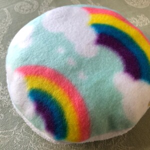 Pode incluir: Um objeto redondo e macio com um fundo azul claro, nuvens brancas e dois arco-íris coloridos. Os arco-íris apresentam faixas rosa, amarelas, azuis e roxas. É provavelmente uma almofada decorativa ou um brinquedo.
