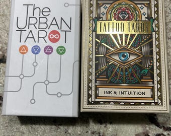 May include: Two tarot card decks. The Urban Tarot deck is white with black text and colorful symbols. The Tattoo Tarot deck is beige with gold accents, a central eye design, and the text "INK & INTUITION."