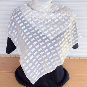Op de afbeelding: Een doorschijnende witte sjaal met een rasterpatroon is over een zwarte top op een mannequin gedrapeerd. Het ontwerp van de sjaal heeft een herhalend patroon van rechthoekige vormen. De achtergrond is een lichtgekleurd oppervlak met lamellen.