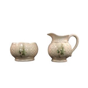 May include: A white ceramic creamer and sugar bowl set with a floral design. The creamer has a handle and a spout. The sugar bowl is round and has a scalloped edge.