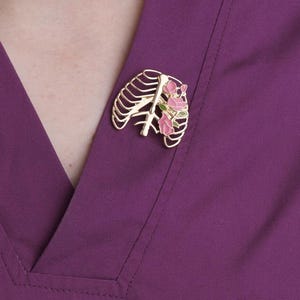 May include: A gold-colored brooch in the shape of a rib cage with pink flowers and green leaves. The brooch is pinned to a purple garment. The design is detailed and the colors are vibrant.