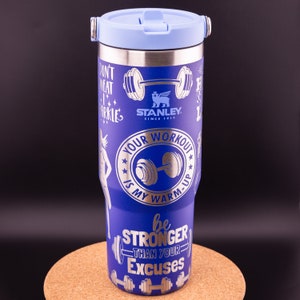 May include: A blue Stanley travel mug with a light blue lid and handle. The mug has silver text and graphics, including the words "Your Workout Is My Warm-Up" and "Be Stronger Than Your Excuses." The Stanley logo is also visible.