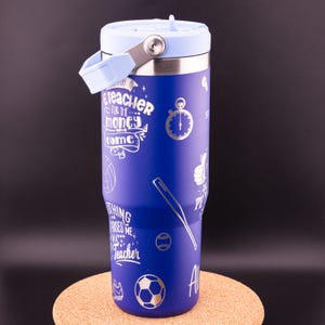 May include: Blue stainless steel tumbler with a light blue lid and handle. The tumbler has a variety of silver-colored designs, including a baseball, basketball, soccer ball, and a baseball bat. The text "I'm a teacher... for the money and fame" is printed on the tumbler.