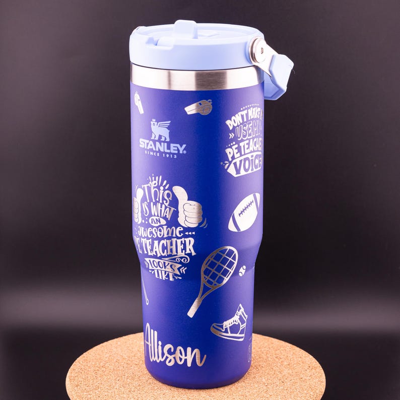 May include: A blue stainless steel travel mug with a light blue lid. The mug has a silver design that says "This is what an awesome teacher looks like" and "Don't make me use my teacher voice". The mug also has a silver design of a whistle, a football, a tennis racket, and a sneaker. The name "Allison" is written in silver at the bottom of the mug.
