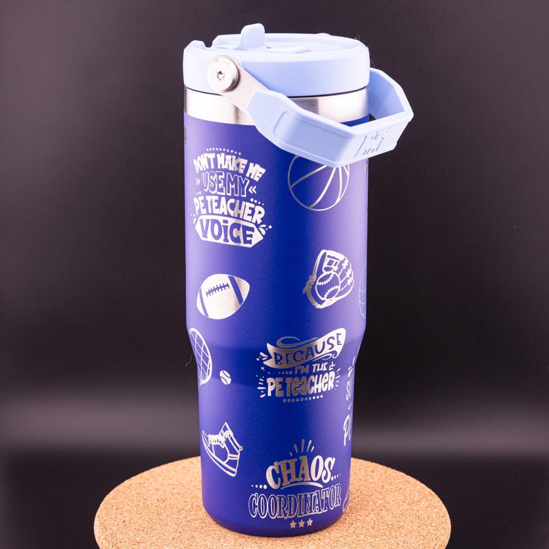 May include: Blue stainless steel tumbler with a silver design. The tumbler has a lid and a handle. The design includes sports-related imagery and the text "Don't make me use my PE teacher voice because I'm the PE teacher... Chaos Coordinator."