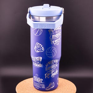 May include: A blue stainless steel tumbler with a light blue lid and a handle. The tumbler has a variety of silver graphics on it, including basketballs, volleyballs, baseballs, and a baseball glove. The graphics also include text that says "PE Teacher", "Chaos Coordinator", and "Nothing Surprises Me, I'm a PE Teacher".