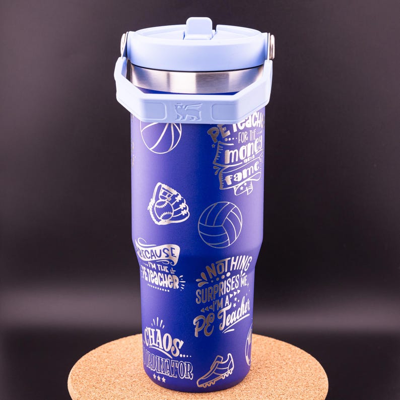 May include: A blue stainless steel tumbler with a light blue lid and a handle. The tumbler has a variety of silver graphics on it, including basketballs, volleyballs, baseballs, and a baseball glove. The graphics also include text that says "PE Teacher", "Chaos Coordinator", and "Nothing Surprises Me, I'm a PE Teacher".