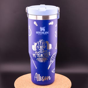 May include: A blue stainless steel travel mug with a light blue lid and a silver handle. The mug has a white design that says "This is what an awesome PE Teacher looks like" with a thumbs up on each side. The mug also has a silver design of a whistle, a stopwatch, a tennis racket, a basketball, and a pair of running shoes. The mug has the name "Allison" printed on the bottom.