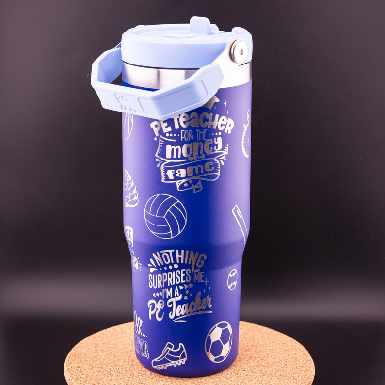 May include: A blue stainless steel tumbler with a light blue lid and handle. It has a silver design with the text "PE Teacher...for the money and fame" and "Nothing surprises me...I'm a PE Teacher".