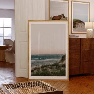 May include: A framed print of a coastal scene with a beach, ocean, and dunes. The large, rectangular print is in a light wood frame and leans against a wall. Two smaller framed prints are visible in the background, one with a horse.
