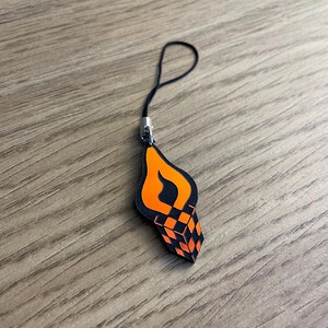 Könnte beinhalten: Ein schwarzer und orangefarbener Handy-Anhänger mit Flammen-Design und einem 3D-Würfelmuster.