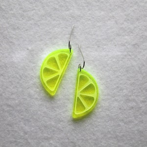 Może przedstawiać: Para neonowo-żółtych kolczyków akrylowych w kształcie ćwiartek limonki. Każdy kolczyk ma srebrny haczyk. Kolczyki to zabawny, letni dodatek.