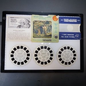 May include: Three View-Master reels with seven three-dimensional full-color pictures each. The reels feature Wild Bill Hickok and Jingles, a Western series. The front of the reels are white with black circular images. The back of the reels are black with white text. The text on the reels reads "Wild Bill Hickok and Jingles" and "The Wells Fargo Robbery".