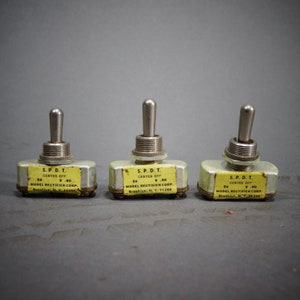 May include: Three vintage S.P.D.T. toggle switches with silver handles and yellow labels. Each switch has the text "S.P.D.T. CENTER OFF" and "MODEL RECTIFIER CORP. Brooklyn, N.Y. 11204" printed on the label. The switches are on a gray background.
