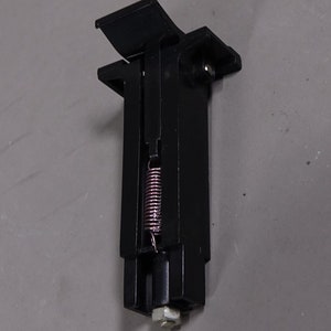 May include: Black metal spring-loaded latch with a metal spring inside. The latch has a rectangular shape with a small arm that extends out from the top.