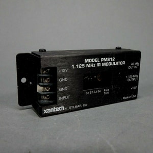 May include: Black box with the text "MODEL PMS12 1.125 MHz IR MODULATOR" and "xantech SYLMAR, CA". The box has several input and output ports labeled with +12V, GND, and INPUT. It also has a frequency trim knob and a switch labeled S1 S2 S3 S4.