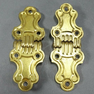 May include: Two antique gold-toned metal hinges with ornate designs. The hinges are closed and have a decorative, ribbed hinge.