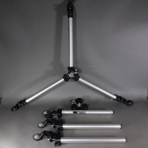 May include: A silver tripod with three legs and a black center. The tripod is disassembled and the legs are laid out on a gray surface. The legs have black wheels on the ends.