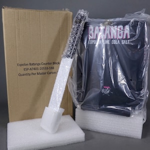 May include: A black counter block with the text "BATANGA ESPOLON LINE COLA SALT" in pink lettering. The block is packaged in plastic wrap and is sitting on a white foam base.