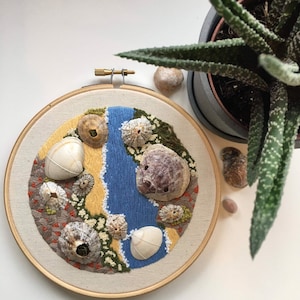May include: A circular embroidery hoop with a beige fabric background. The embroidery depicts a blue river flowing through a beach scene with various seashells and a yellow sand area. The embroidery is surrounded by a wooden hoop.