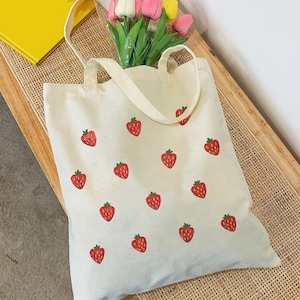 Może przedstawiać: Biała torba typu tote z płótna z wzorem czerwonych truskawek. Torba ma dwa uchwyty i jest wykonana z lekkiej tkaniny.