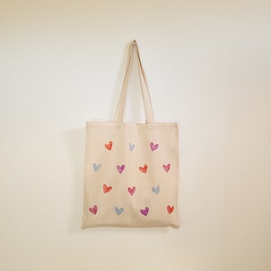 May include: A beige tote bag with a design of colorful hearts printed on it. The bag has two handles and is hanging on a white wall.