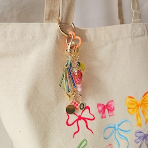 May include: A gold key chain with a heart charm, beads, and a strawberry charm. The key chain is attached to a white tote bag with a pink, yellow, and blue bow design.
